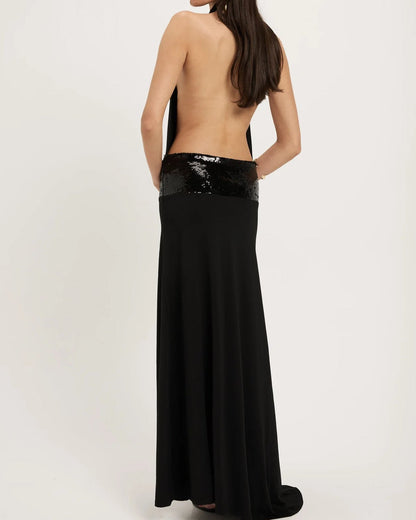 The Ance Gria Adele Gown in Black is an elegant floor-length gown featuring a halter neckline with a softly draped cowl front that creates a sophisticated silhouette. The open back adds a striking statement, while a low-sitting sequinned waistband accentuates the hips with subtle sparkle. Crafted in classic black, this designer gown is perfect for black-tie events, formal occasions., gala dinners, weddings, and evening celebration, combining timeless elegance with modern glamour.