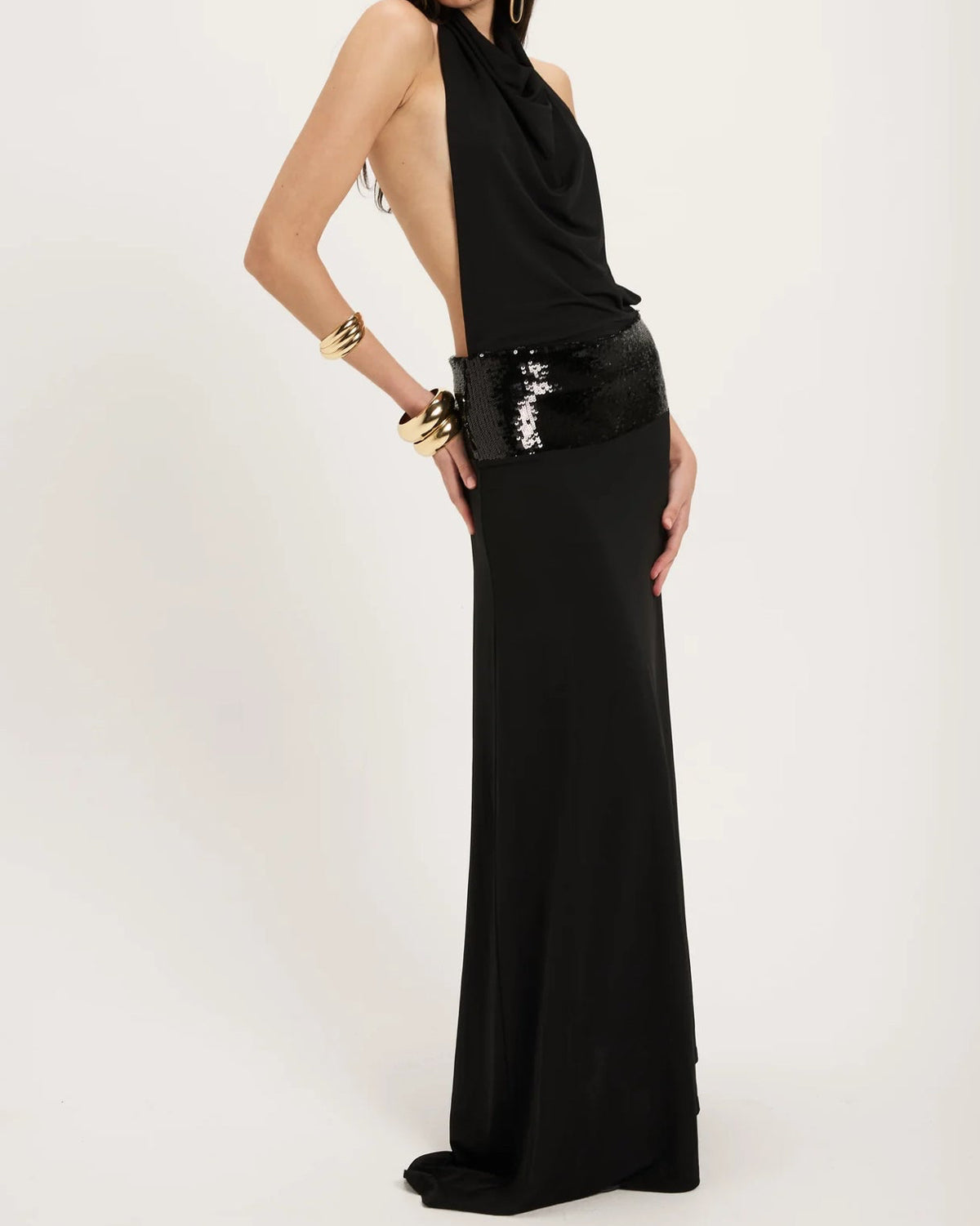 The Ance Gria Adele Gown in Black is an elegant floor-length gown featuring a halter neckline with a softly draped cowl front that creates a sophisticated silhouette. The open back adds a striking statement, while a low-sitting sequinned waistband accentuates the hips with subtle sparkle. Crafted in classic black, this designer gown is perfect for black-tie events, formal occasions., gala dinners, weddings, and evening celebration, combining timeless elegance with modern glamour.