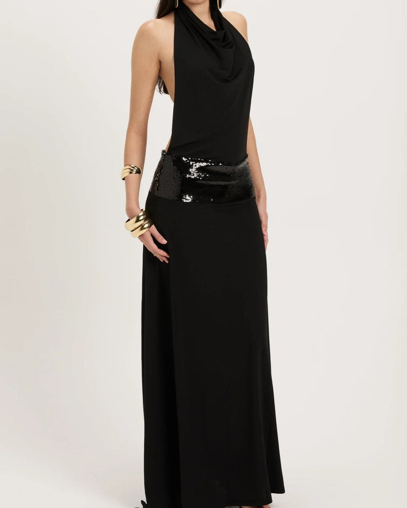 The Ance Gria Adele Gown in Black is an elegant floor-length gown featuring a halter neckline with a softly draped cowl front that creates a sophisticated silhouette. The open back adds a striking statement, while a low-sitting sequinned waistband accentuates the hips with subtle sparkle. Crafted in classic black, this designer gown is perfect for black-tie events, formal occasions., gala dinners, weddings, and evening celebration, combining timeless elegance with modern glamour.