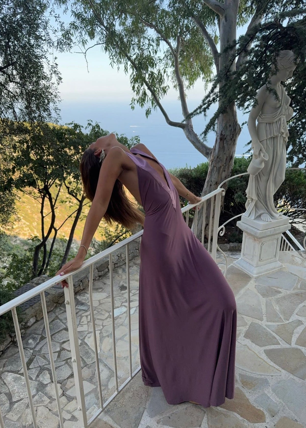 Model wearing the Ance Gria Rosa Maxi Dress in Mauve, featuring a plunging V-neckline, wide shoulder straps, a fitted waist and a soft floor-length skirt with an elegant draped silhouette. Back view of the Ance Gria Rosa Maxi Dress in Mauve, showcasing a dramatic low open V-back with wide shoulder straps and a flowing floor-length skirt in soft stretch fabric.