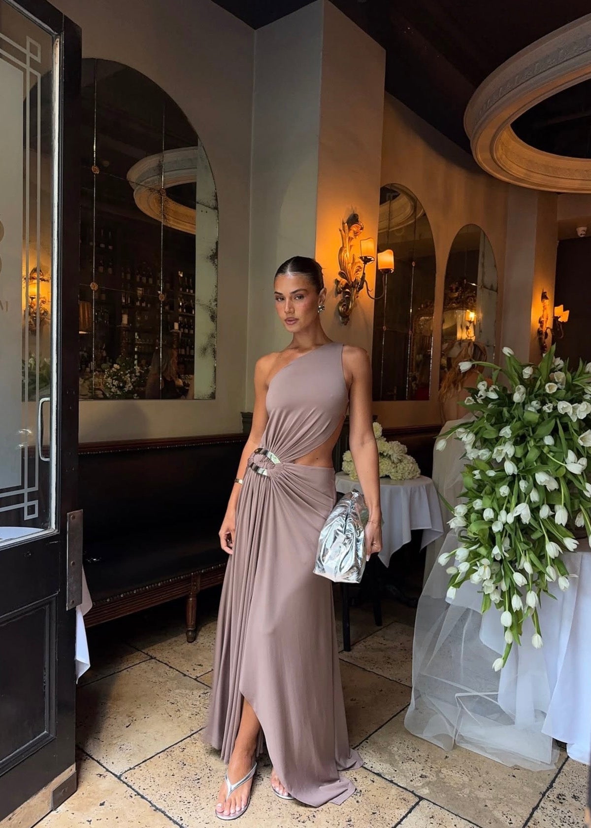 Model wearing the Ance Gria Serena Maxi Dress in Taupe, designed with a one-shoulder bodice, soft draping, gathered waist with a statement ring detail, side cut-out and an elegant floor-length skirt with an asymmetrical hem.