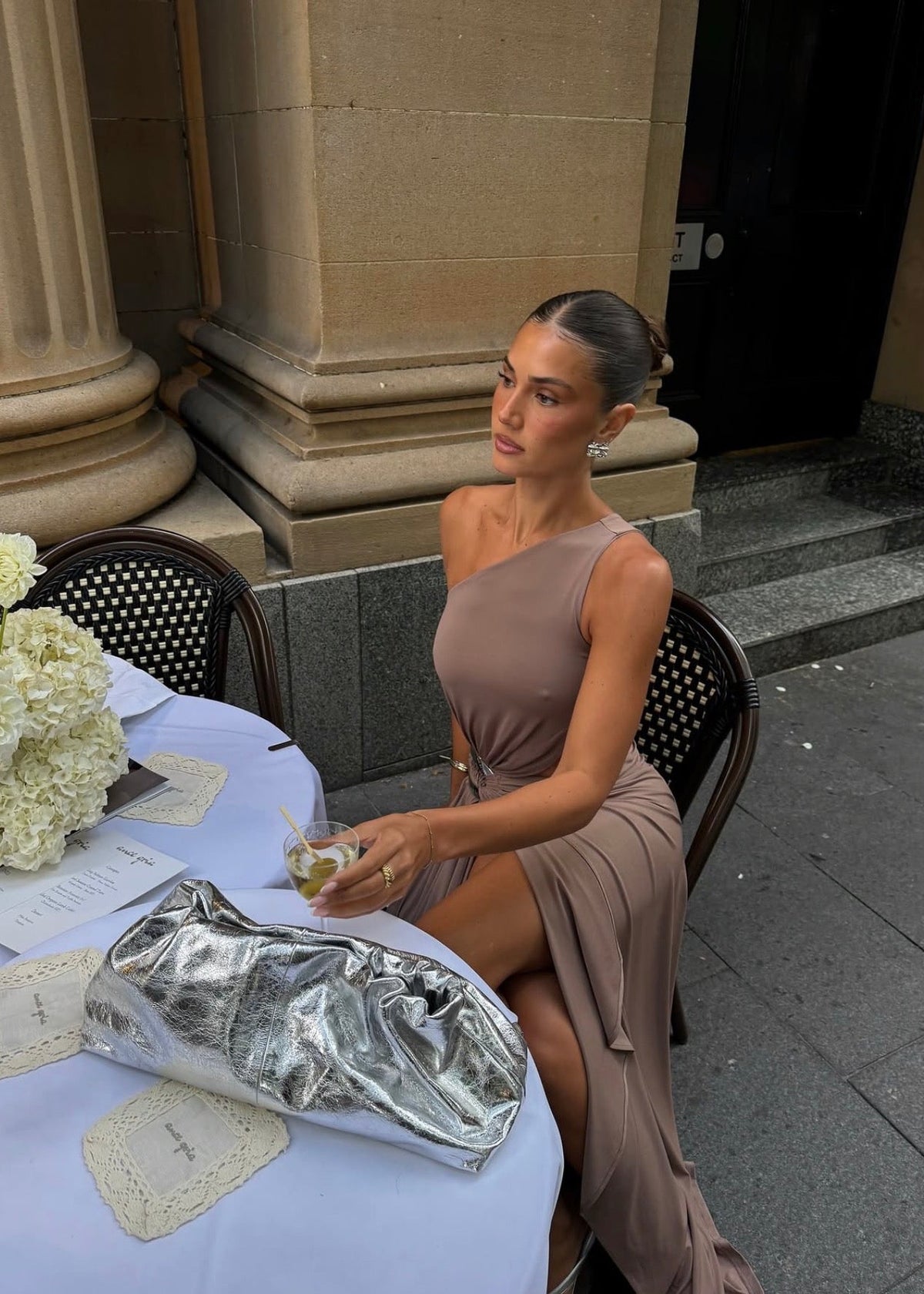 Model wearing the Ance Gria Serena Maxi Dress in Taupe, designed with a one-shoulder bodice, soft draping, gathered waist with a statement ring detail, side cut-out and an elegant floor-length skirt with an asymmetrical hem.