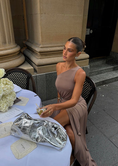 Model wearing the Ance Gria Serena Maxi Dress in Taupe, designed with a one-shoulder bodice, soft draping, gathered waist with a statement ring detail, side cut-out and an elegant floor-length skirt with an asymmetrical hem.