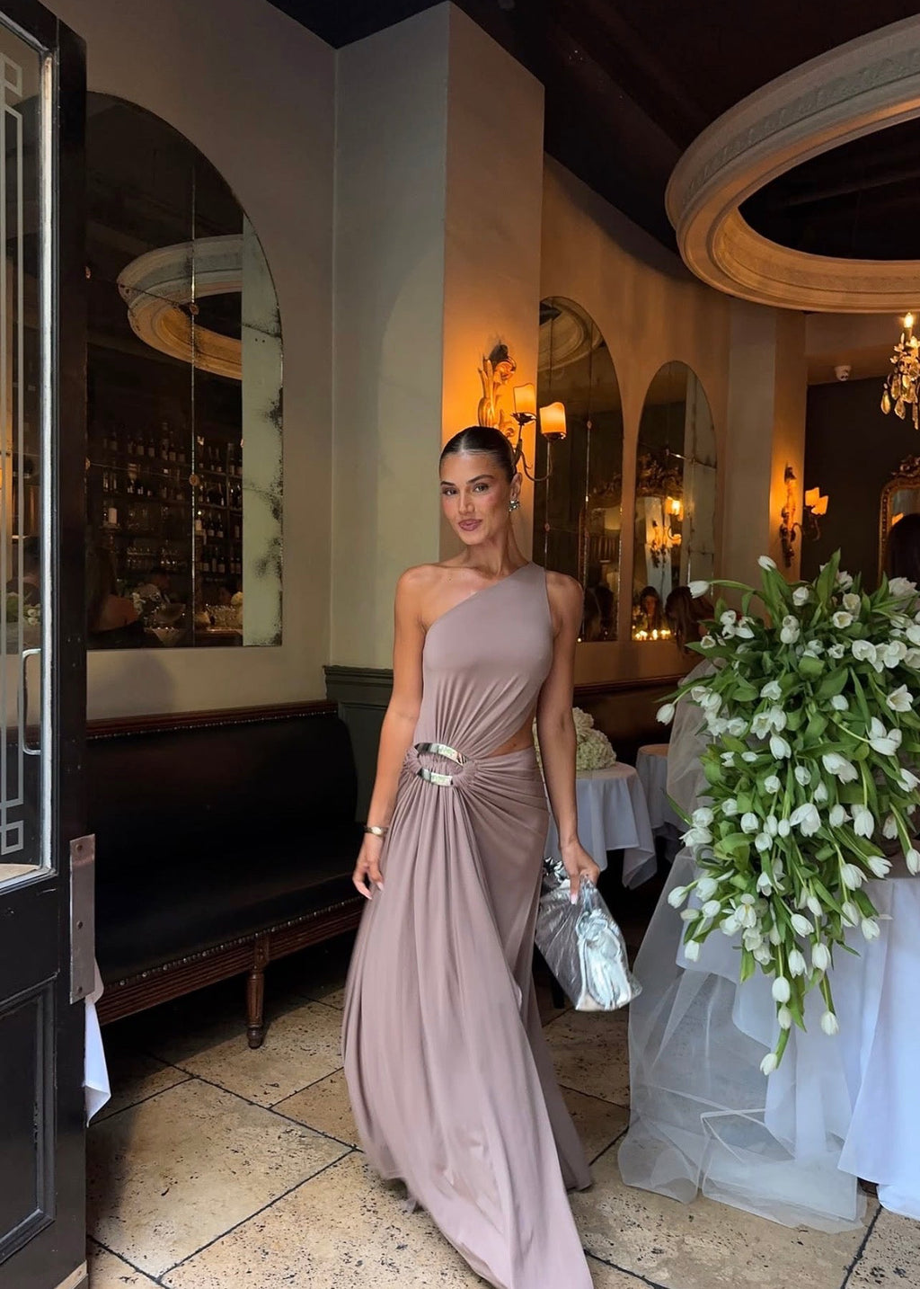 Model wearing the Ance Gria Serena Maxi Dress in Taupe, designed with a one-shoulder bodice, soft draping, gathered waist with a statement ring detail, side cut-out and an elegant floor-length skirt with an asymmetrical hem.