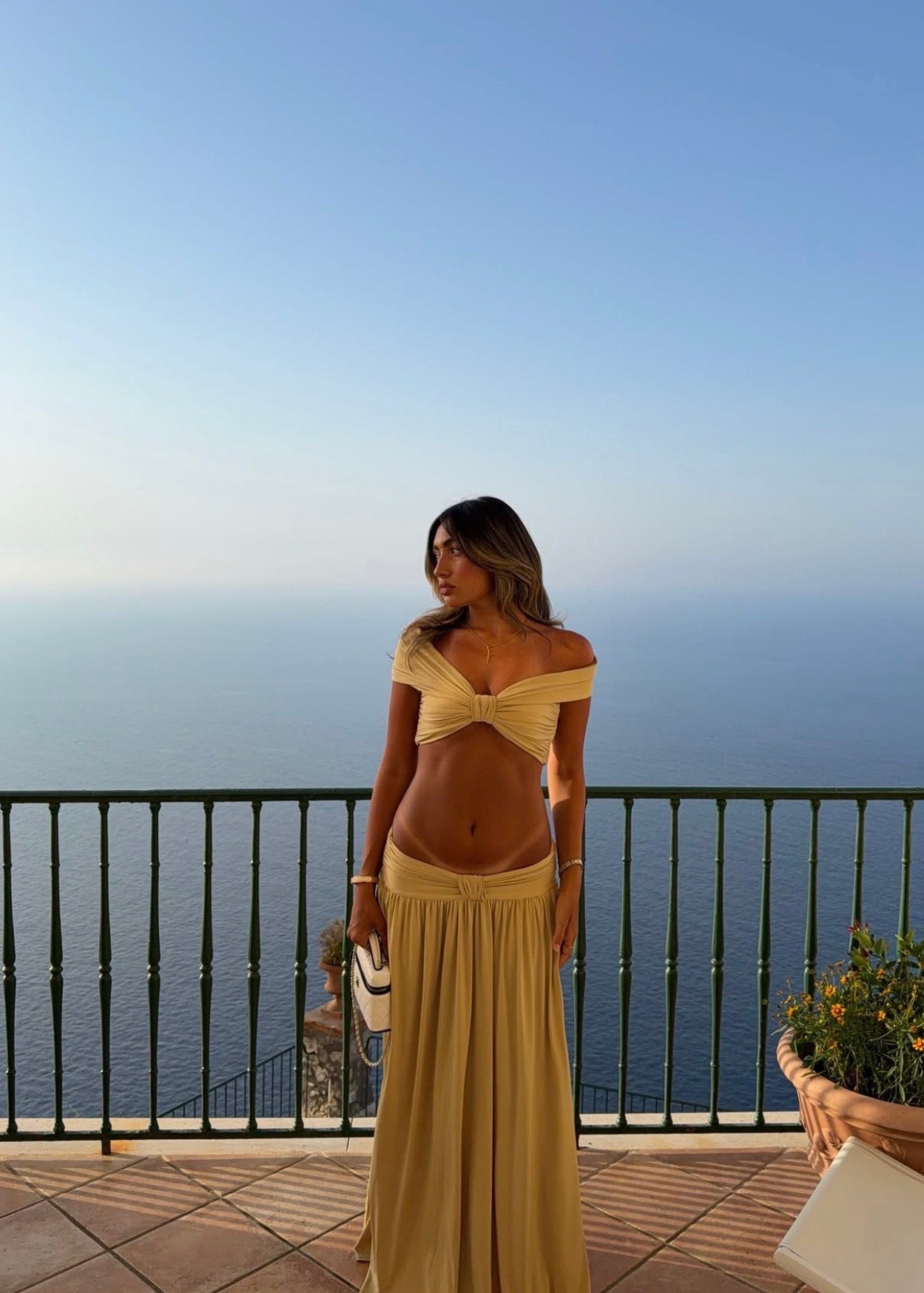 Model wearing the Arcina Ori Alessia Set in Sand, featuring an off-the-shoulder twist-front crop top with draped short sleeves and gathered bust detailing, paired with a low-rise maxi skirt with a gathered waistband, soft draping and a floor-length flowing silhouette.