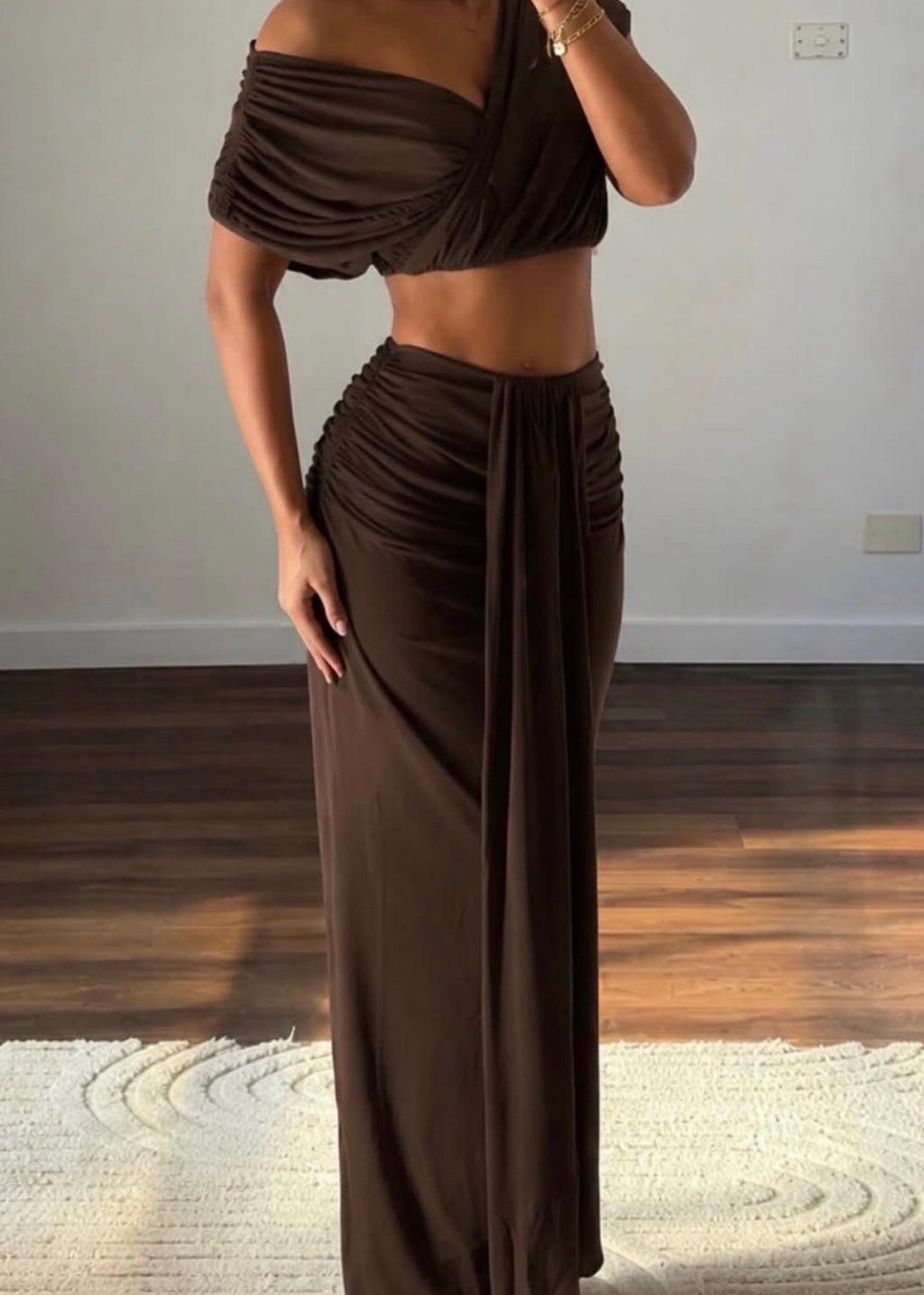 Model wearing the Benni Celia Set in Chocolate, featuring an off-the-shoulder draped crop top with a crossover neckline, slim shoulder straps and a matching high-waisted maxi skirt with gathered detailing, a front draped panel and a flowing full-length silhouette.